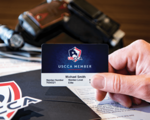 A Caucasian hand holding a blue USCCA Membership card. On the tabletop below is the folder of the USCCA welcome package as well as a holstered pistol.