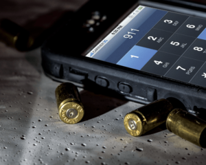 A black smartphone's keypad has been used to dial 911. The phone lies next to three spent 9mm brass casings.