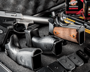 A selection of semi-automatic pistols with their muzzles inserted into the protective foam of a hard-sided case. A row of black magazines show the Ruger logo on the butt plate and a few boxes of SIG and Black Hills ammunition are stacked nearby. A silver and black Ruger pistol lies beside the others, uncased.