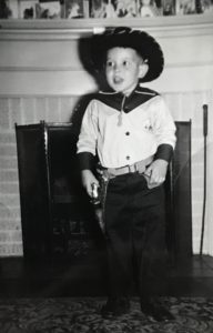 Black and white vintage photograph of a young boy dressed up in a cowboy shirt, black hat and a gunbelt with a six-shooter cap gun, standing before a mantle in someone's living room.