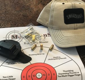 A tan baseball cap with a black Walther patch on the front, a black inside-the-waistband (IWB) holster, a clear bag contaning a pair of bright yellow foam earplugs and three rounds of .380 wadcutter pistol ammunition lie atop a right-hand shooter aim correction paper target with a red bullseye.