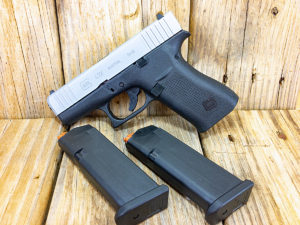 Two-tone silver and black Glock 43X subcompact pistol propped against a backdrop of unstained wood. Two empty Glock magazines lie nearby.