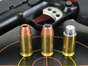 Three rounds of ammo standng atop a target with a handgun lying nearby in the background. The three cartridges have different bullet weights and therefore different velocities.