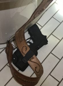 A black pistol creeping out of a black OWB holster attached to a brown leather western-style belt. The rig is lying on the tiled floor of a bathroom stall with the belt snaking up the side of the toilet.